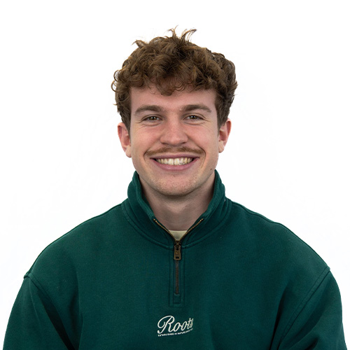 Young man with curly brown hair smiling, wearing a green quarter-zip fleece with a Roots logo on the chest.