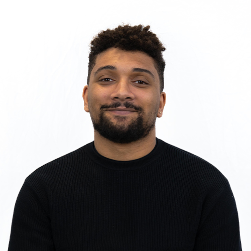 Portrait of a man with short curly hair and a beard, wearing a black sweater, smiling at the camera against a white background.