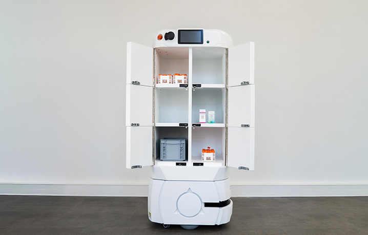 White autonomous medication-dispensing robot with an open cabinet showing medicine bottles on shelves in a clinic setting.
