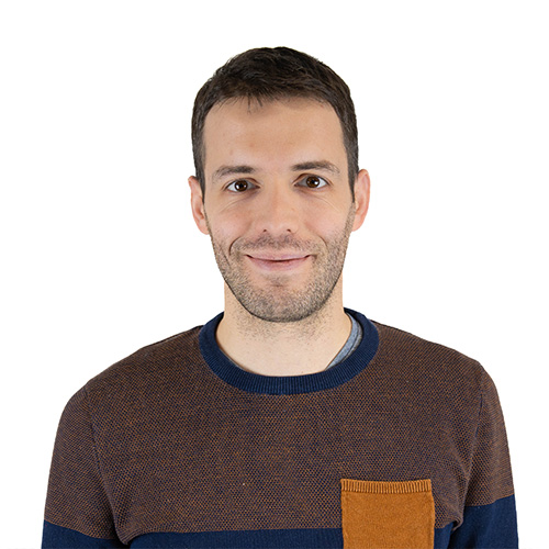 Portrait of a man with short dark hair and stubble, wearing a brown sweater with a contrasting orange pocket, smiling at the camera