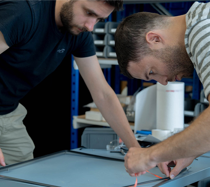 Two men in a workshop lean over a metal frame, wiring an orange cable.