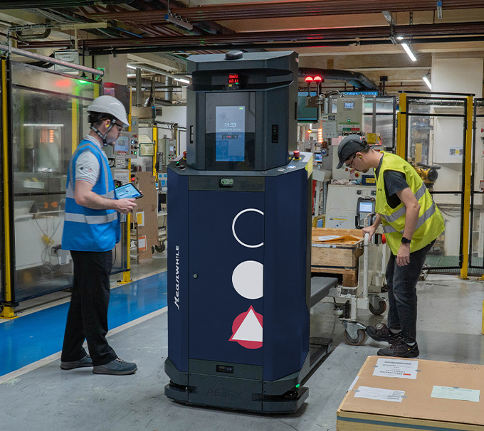 Factory floor with a blue autonomous robot between two workers in safety vests and hard hats.