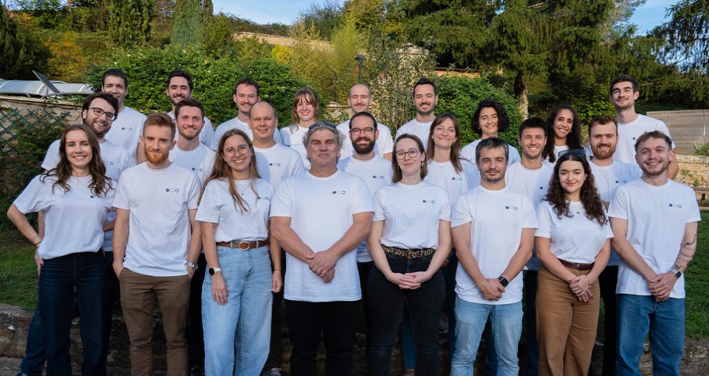 Group of smiling people wearing white shirts posing outdoors for a team photo, arranged in rows on a sunny day