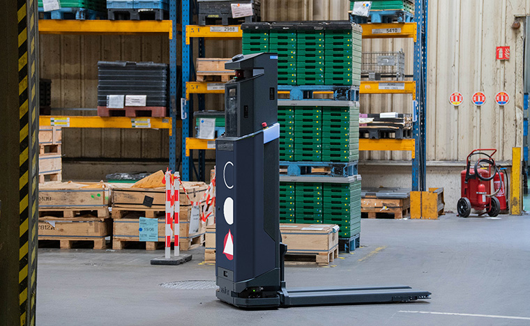 Autonomous pallet-moving robot in a warehouse corridor beside stacked pallets and shelving