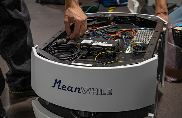 Close-up of a circular service robot with exposed internal wiring and a technician adjusting cables; the front reads 'MeanWhile'.