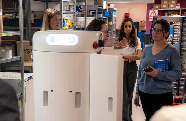 A group of four women in a warehouse discussing a tall white service robot during a demonstration.