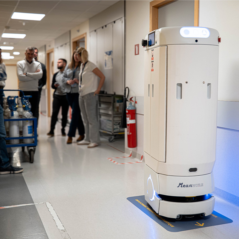 White autonomous hospital robot on a blue mat with arrows in a hallway, while a group of staff stand and chat in the background.