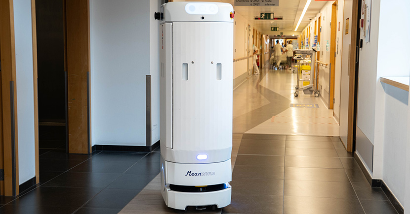 White autonomous hospital service robot moving down a hallway, with staff and carts visible in the background.