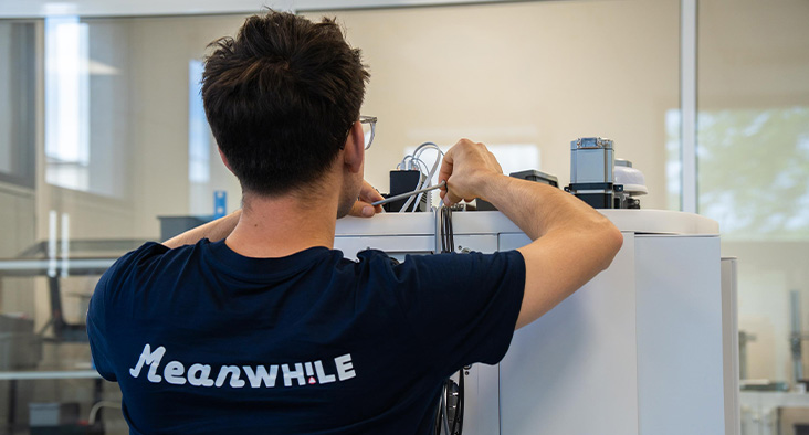 Back view of a technician in a navy shirt with 'Meanwhile' connecting cables to a white laboratory instrument in a bright lab.
