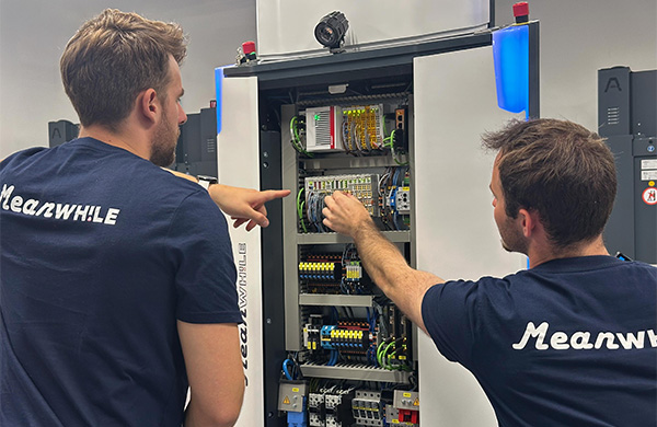 Two technicians in matching blue shirts inspect the open industrial control cabinet, pointing at wiring inside a workshop setting.