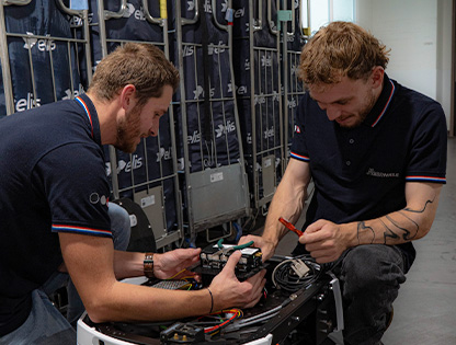Two technicians work on a circuit board inside a data center, using tools and cables.