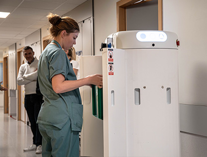 Nurse in green scrubs loads a medical device in a hospital corridor with another staff member in the background.