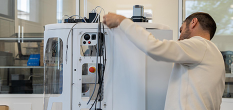 Technician in a white coat adjusts a large white laboratory instrument, routing cables and inspecting its controls.