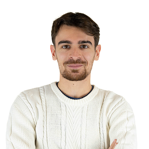 Portrait of a man with short brown hair and light stubble, wearing a white knitted sweater and a subtle smile.