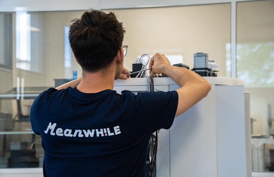 Person in a dark blue shirt with 'MEANWHILE' on the back wires a large lab device, seen from behind, in a bright lab setting